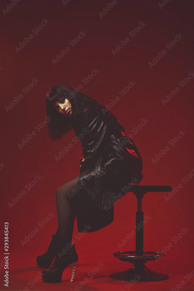 Attractive young woman in black leather coat and stockings on black shoes alluring posing. Fashion shoot in red studio. Sexy poster with pretty brunette girl. female sitting on chair