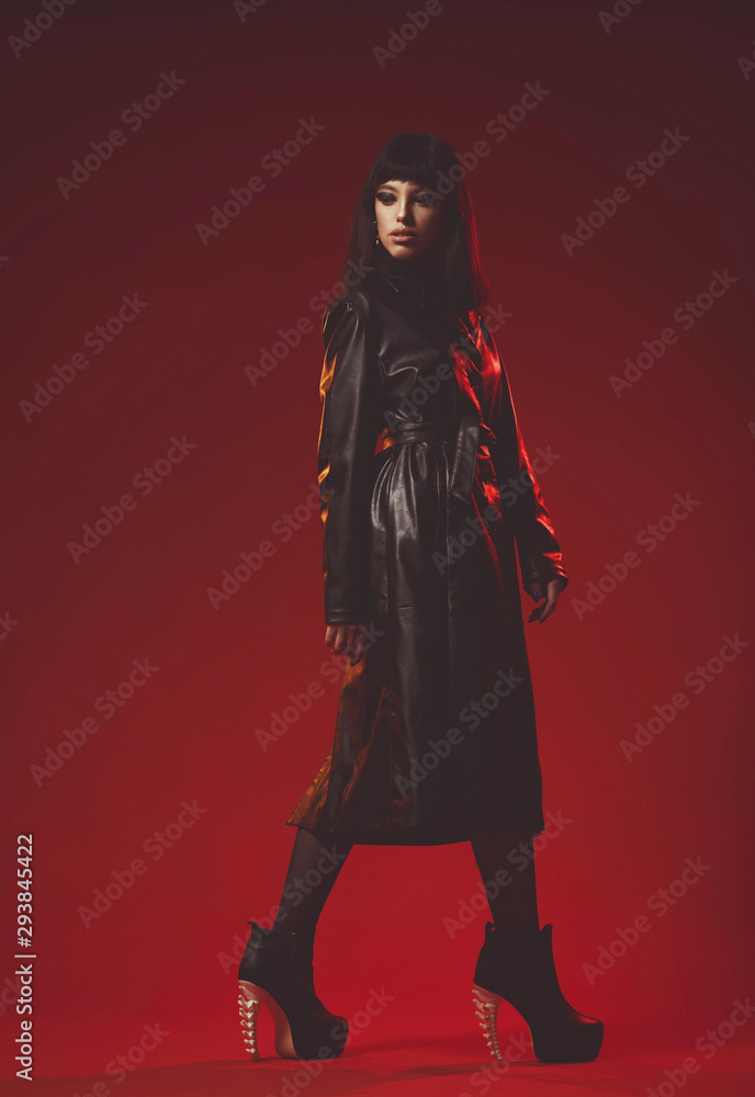 Attractive young woman in black leather coat and stockings on black shoes alluring posing. Fashion shoot in red studio. Sexy poster with pretty brunette girl.
