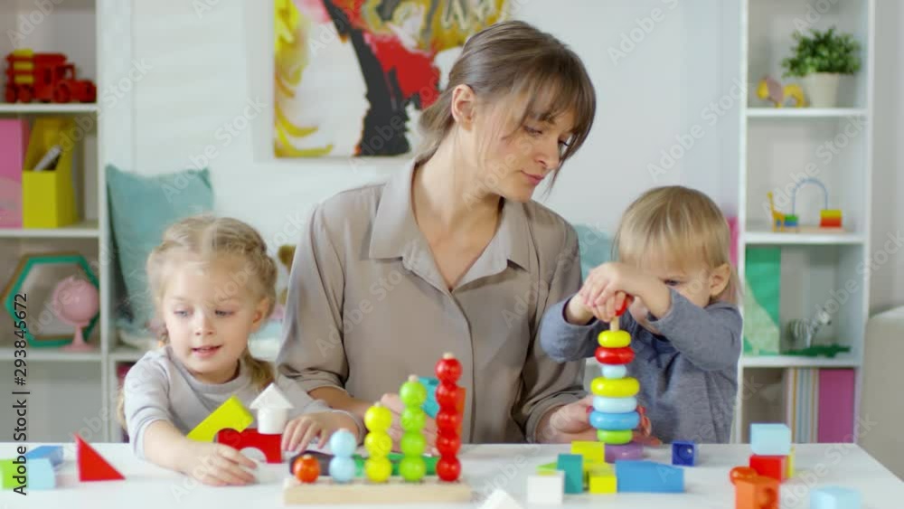 Mother and little daughter and son playing with educational toys and speaking while sitting together at table in kids room Mother and little daughter and son playing with educational toys and speaking while sitting together at table in kids room
