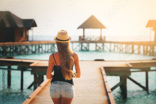 Summer vacation holiday travel tropical beach background - beautiful young Caucasian girl tourist walking on the pier in lagoon at sunset with hat near the sea on the exotic island