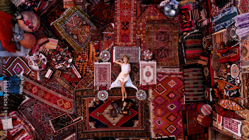Obraz premium Beautiful happy girl in a long white dress laying on the carpet and rugs in Goreme, Cappadocia, Turkey. Top view drone.