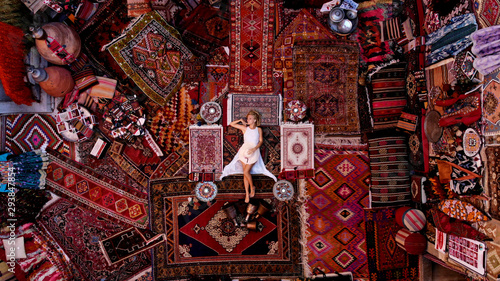 Beautiful happy girl in a long white dress laying on the carpet and rugs in Goreme, Cappadocia, Turkey. Top view drone.