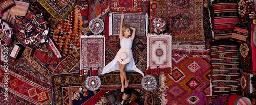 Beautiful happy girl in a long white dress laying on the carpet and rugs in Goreme, Cappadocia, Turkey. Top view drone.