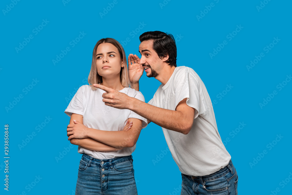 Bearded man with mustaches in white t-shirt tells secret to a young blond girl in white t-shirts and blue jeans standing isolated over blue background. Woman looks surprised. Secret telling. Bearded man with mustaches in white t-shirt tells secret to a young blond girl in white t-shirts and blue jeans standing isolated over blue background. Woman looks surprised. Secret telling.
