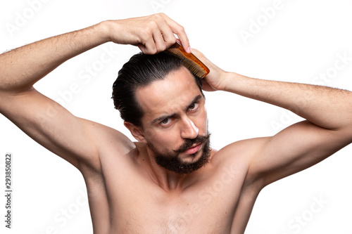 Stylish handsome bearded with mustaches combing hair standning bare isolated over white background. Concept of morning treatment. Morning routine