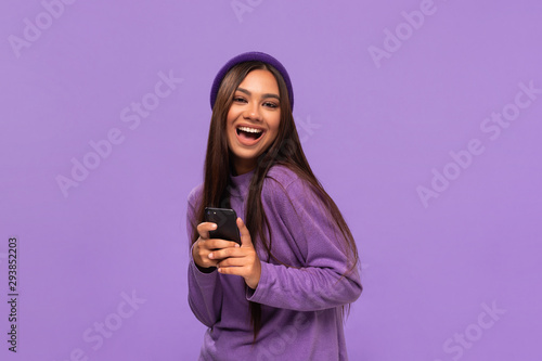 Smiling african-american girl in a hat and sweater holding smartphone standing isolated over purple background.