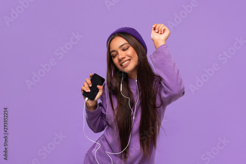 Pretty african-american girl in a hat and sweater listening to music on a mobile phone with wired headphones and dancing isolated over purple background. Enjoying life.