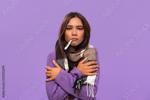 Pretty african-american girl in a sweater and cosy checked scarf checking thermometer standing isolated over purple background. Catching cold in winter.