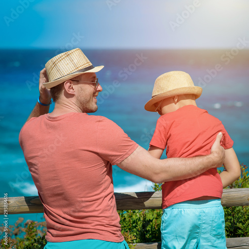Cute European boy is on his father’s hands in front of picturesque wavy sea, enjoying summer holidays. They wearing similar clothes sun hats and sunglasses smiling and looking to each other. © Artem