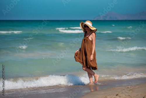 Young European lady in colorful bonnet hat and beautiful dress walking along picturesque wavy sea, she enjoying serenity around. photo