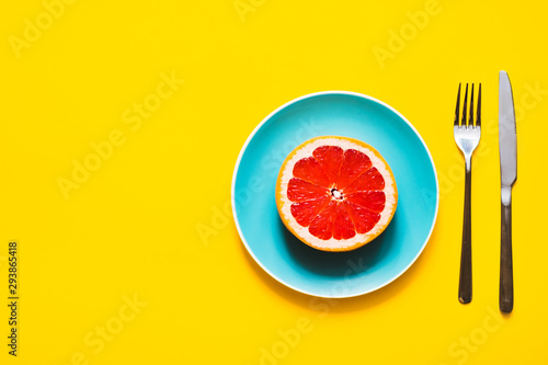 Concept of vegan diet. High angle shot - Appetizing juicy fresh grapefruit on color plate with fork and knife on yellow background with copy space.