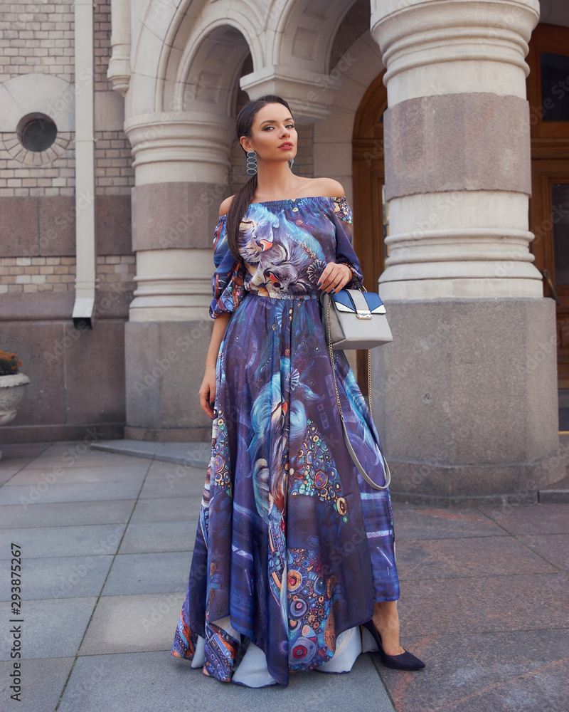 Young beautiful brunette woman with long straight hair in a tail wearing colorful blue dress, holding gray leather handbag and walking old city street