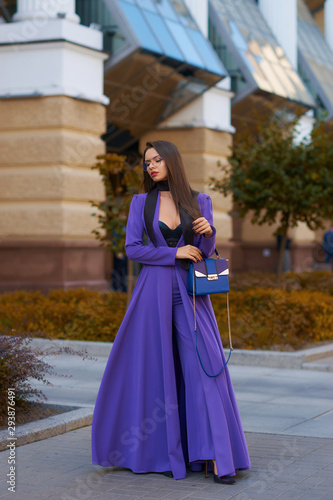 Young beautiful caucasian woman with long straight hair and makeup in purple trousers and long coat cape standing and posing at city street in autumn © Dmitry Tsvetkov