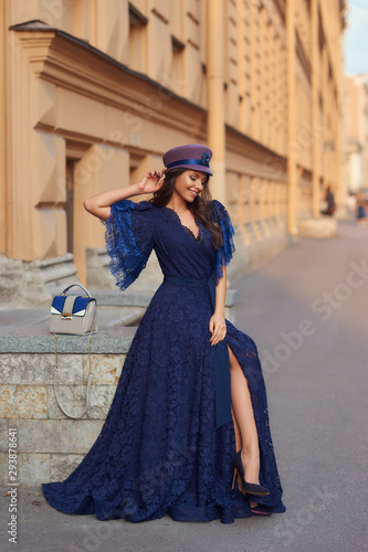 Full length outdoor portrait of young beautiful elegant woman in long blue evening dress and purple hat with gray leather handbag sitting and posing at old city street on a sunny evening day © Dmitry Tsvetkov