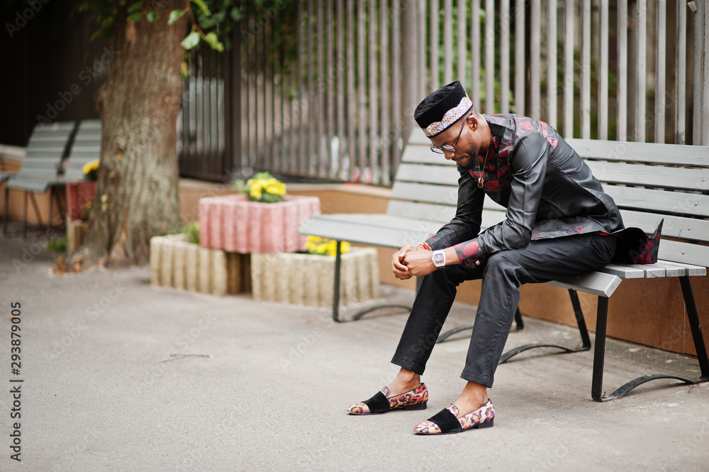 Handsome afro american man wearing traditional clothes, cap and eyeglasses in modern city sitting on bench.