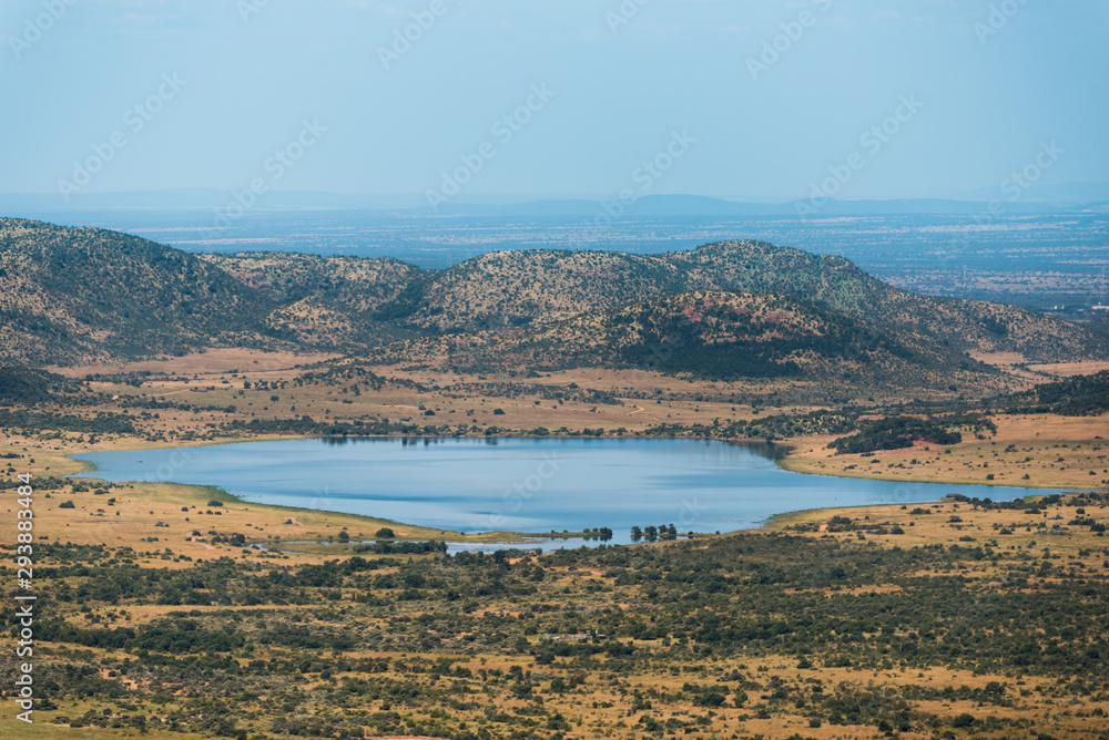 Obraz premium A dam/lake at the foot of the hills, surrounded by the dry and bushy plains of Pilanesberg Nature Reserve in South Africa