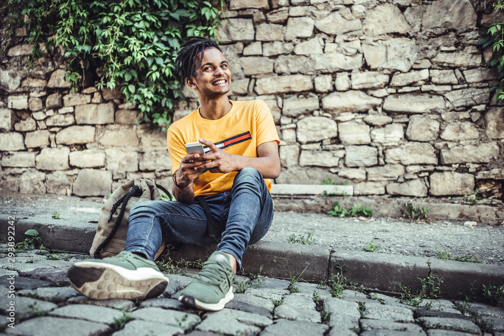 Fototapeta premium Happy student sitting near stone wall and using mobile phone