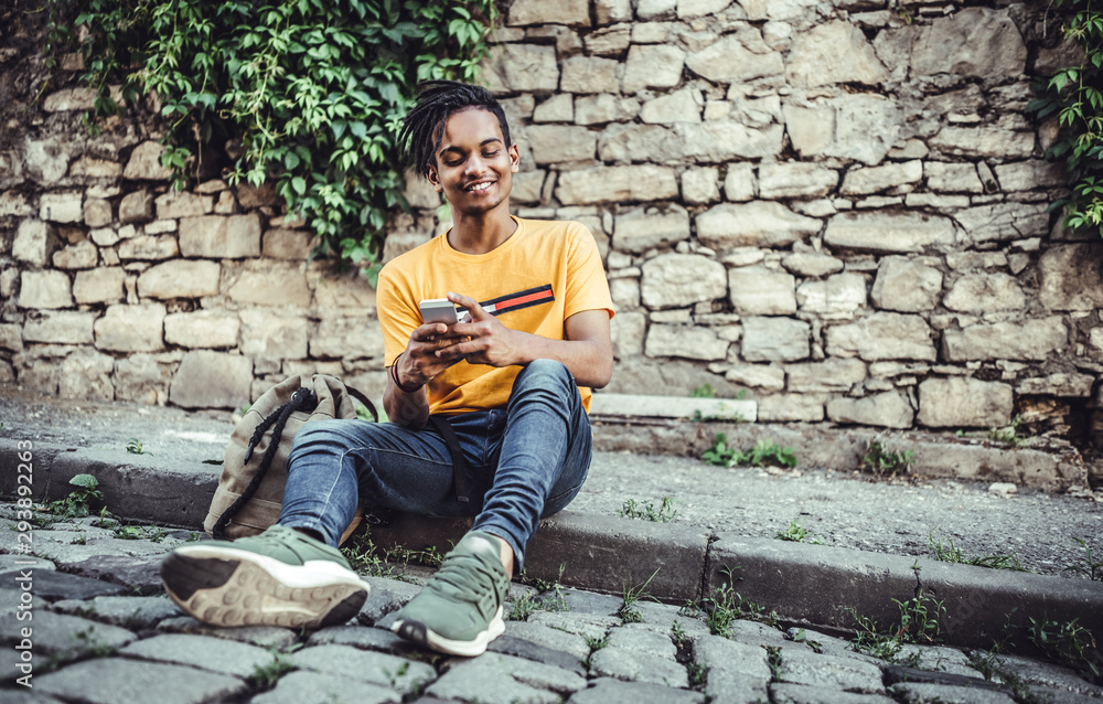 Fototapeta premium Handsome young man with backpack sitting near stone wall and using cell phone in the city outdoors
