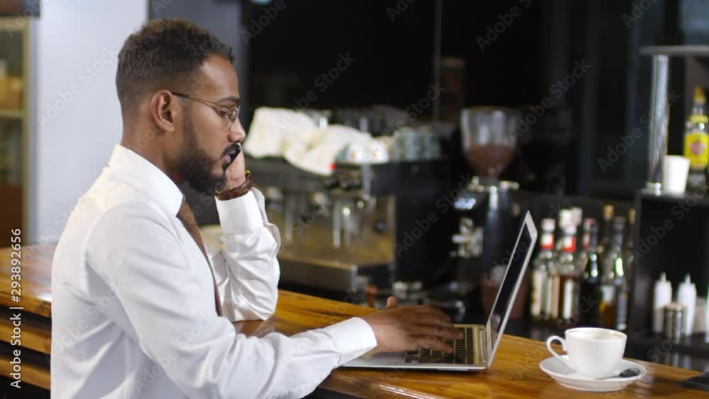 Middle eastern businessman in formal shirt and tie typing on laptop and talking on mobile phone while working at bar counter in cafe Middle eastern businessman in formal shirt and tie typing on laptop and talking on mobile phone while working at bar counter in cafe