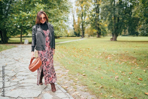 Smiling female dressed boho fashion style colorful long dress with black leather biker jacket with brown leather flap bag having a autumn park walking.