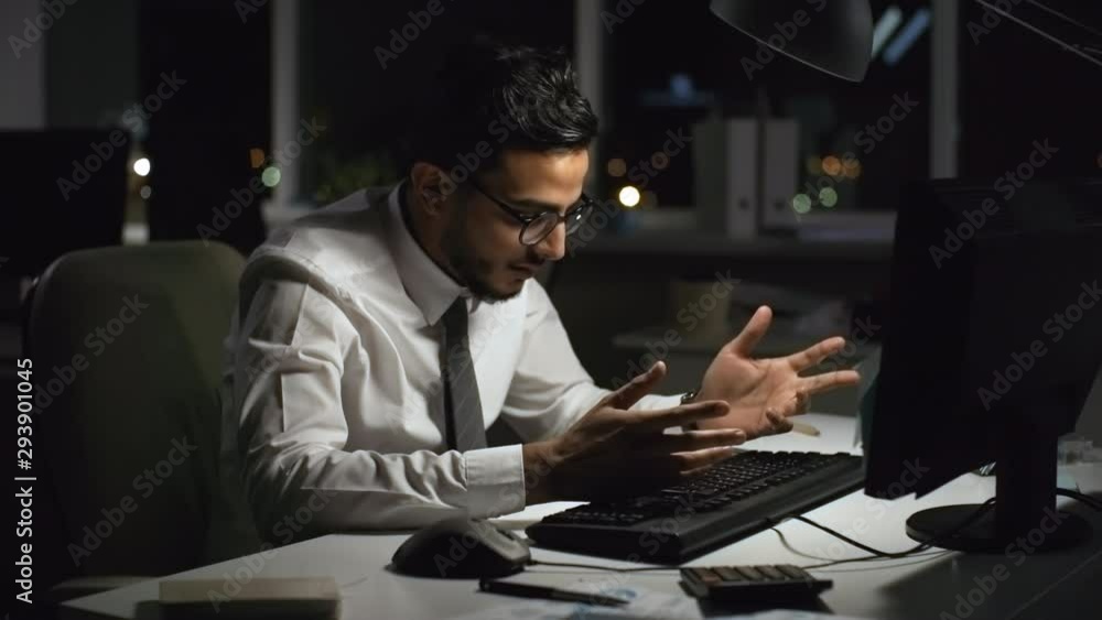 Medium shot of exhausted Arab businessman working on computer in office at night, then hitting keyboard in frustration Medium shot of exhausted Arab businessman working on computer in office at night, then hitting keyboard in frustration