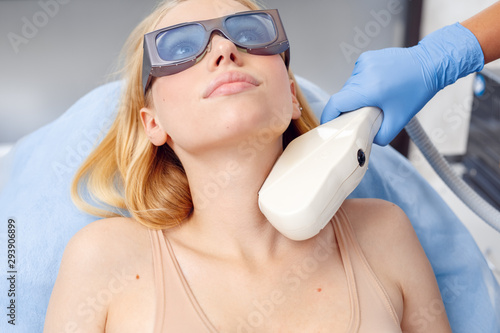 Cosmetology Service. Young woman at beauty clinic lying on medical bed in safety goggles pensive while doctor using photorejuvenation laser on her neck