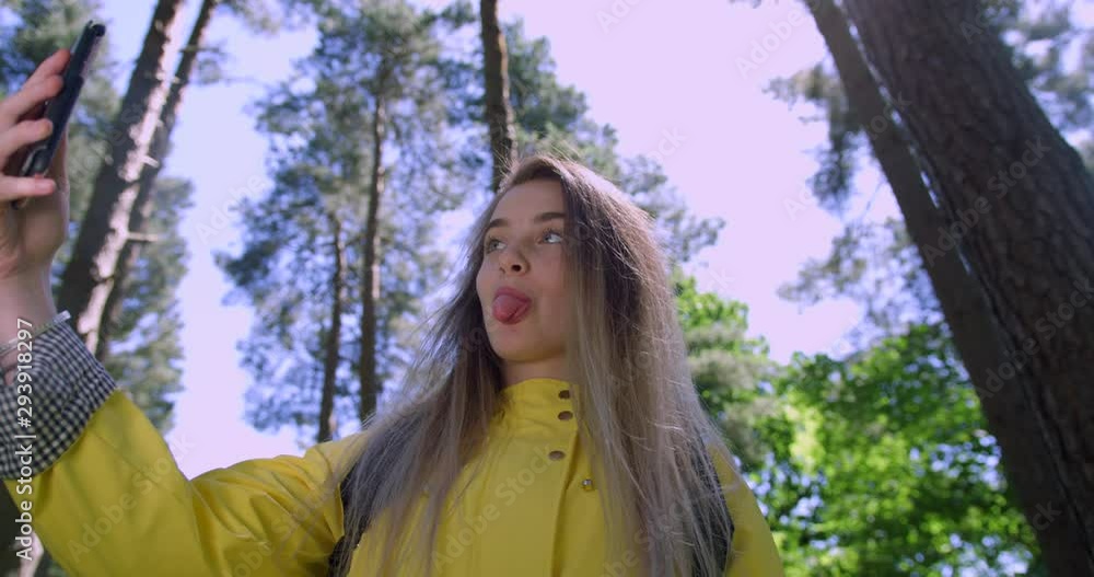 Young Smiling Woman taking a Funny Face Selfie with Mobile Phone in the Forest. Girl in Woodland Glade wearing a Yellow Rain Coat. Happy, blonde Student Girl her Cell within Tree foliage Young Smiling Woman taking a Funny Face Selfie with Mobile Phone in the Forest. Girl in Woodland Glade wearing a Yellow Rain Coat. Happy, blonde Student Girl her Cell within Tree foliage