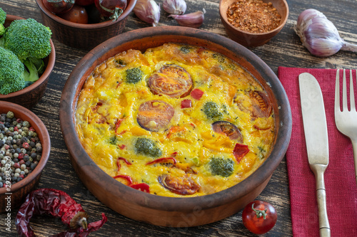 Ceramic bowl with vegetable frittata, simple vegetarian food. Frittata with egg, tomato, pepper, onion, broccoli and cheese on wooden table. Italian egg omelette