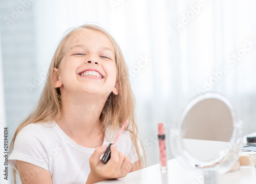 Happy young girl puts lipstick on her lips looking in the mirror at home