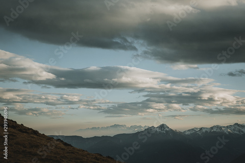 Panorama of snowy peaks of the mountains with beautiful cloud sky above them during orange sunset with haze in the valley with yellow blurry meadow in front