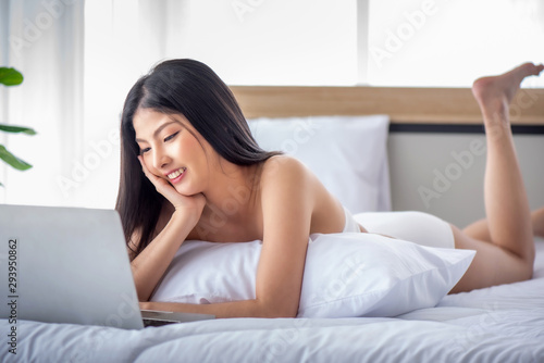 Pretty attractive sensuality young Asian woman wearing red bra and using laptop, laying on the white bed at bedroom in the morning