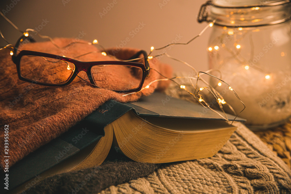 Cozy warmly lit reading area or nook for winter and fall weather with led string lights, books, glasses, wicker table and wool blanket or throw - obrazy, fototapety, plakaty 