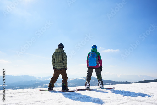 Back view of two tourists snowboarders in sportive equipment standing on snowboards on copy space background of blue sky and woody mountains on sunny winter day. Extreme winter sports concept.