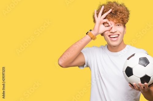 Young handsome man holding soccer football ball with happy face smiling doing ok sign with hand on eye looking through fingers
