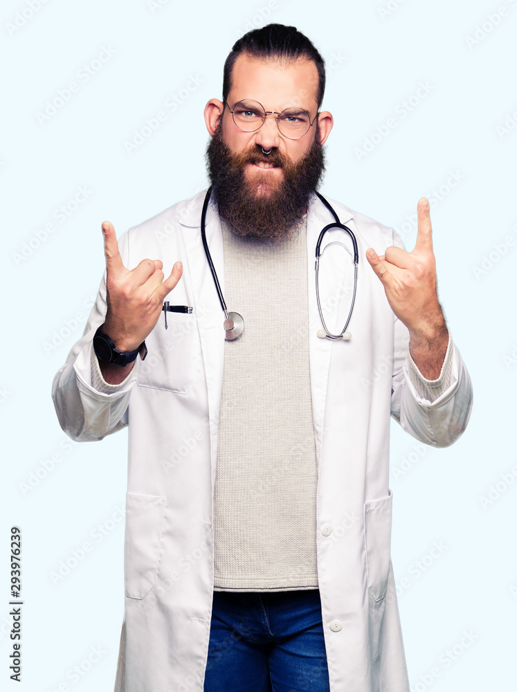 Young blond doctor man with beard wearing medical coat shouting with crazy expression doing rock symbol with hands up. Music star. Heavy concept. Young blond doctor man with beard wearing medical coat shouting with crazy expression doing rock symbol with hands up. Music star. Heavy concept.