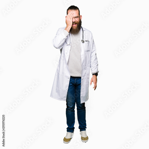 Young blond doctor man with beard wearing medical coat covering one eye with hand with confident smile on face and surprise emotion.