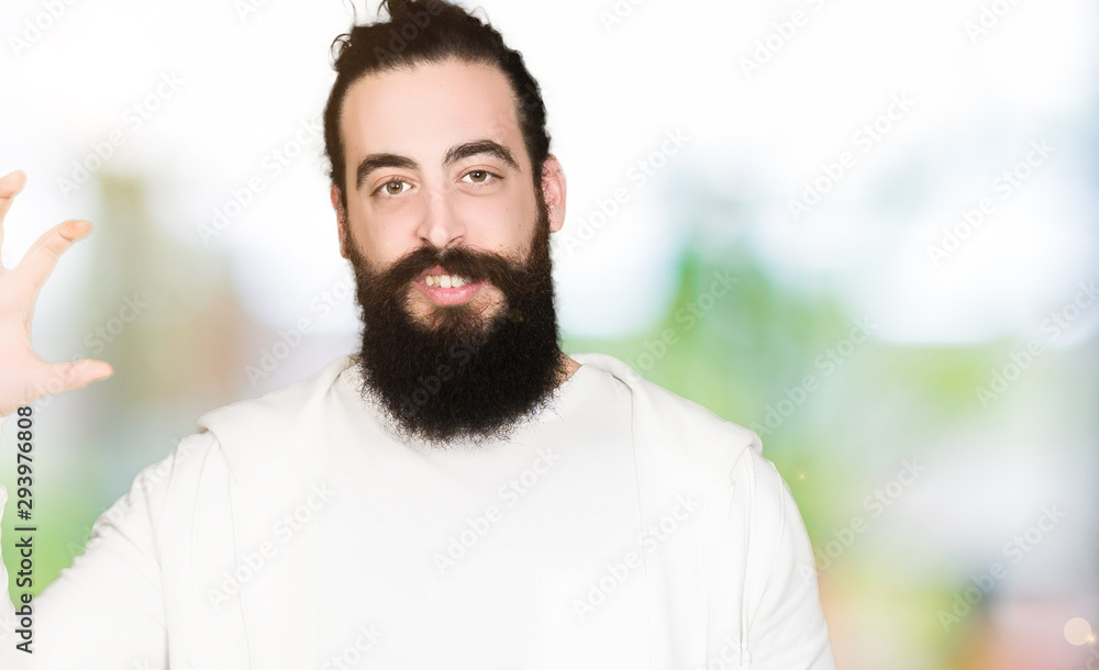 Young man with long hair and beard wearing sporty sweatshirt smiling and confident gesturing with hand doing size sign with fingers while looking and the camera. Measure concept. Young man with long hair and beard wearing sporty sweatshirt smiling and confident gesturing with hand doing size sign with fingers while looking and the camera. Measure concept.