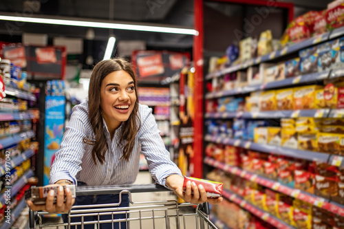 Smiling woman at supermarket. Happy woman at supermarket. Beautiful young woman shopping in a grocery store/supermarket. Shopaholic woman enjoying shopping spree in supermarket