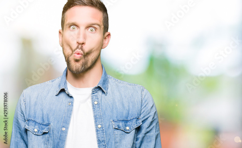 Handsome man with blue eyes and beard wearing denim jacket making fish face with lips, crazy and comical gesture. Funny expression.
