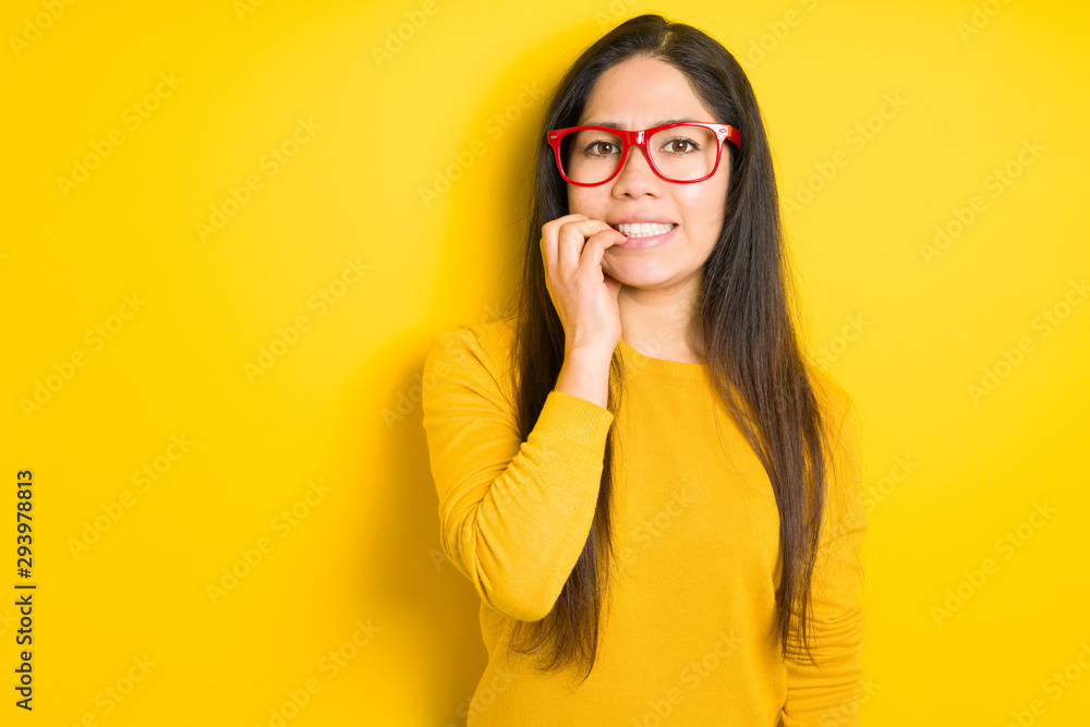 Beautiful brunette woman wearing red glasses over yellow isolated background looking stressed and nervous with hands on mouth biting nails. Anxiety problem.