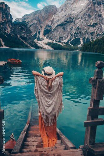 Girl in a straw hat on the background of the turquoise lake with wooden boats in mountains. Safari style blonde woman back view. Dolomites Alps  lago di Braies  Italy