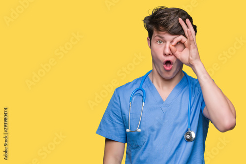 Young doctor wearing medical uniform over isolated background doing ok gesture shocked with surprised face, eye looking through fingers. Unbelieving expression.