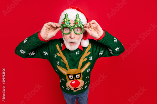 Photo of funny grey haired santa looking on unbelievable low x-mas shopping prices speechless wear x-mas tree shape specs ugly ornament sweater isolated red background