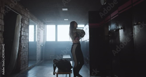 Beautiful young happy Caucasian woman changing clothes after gym exercise in dark locker room, life motivation concept.