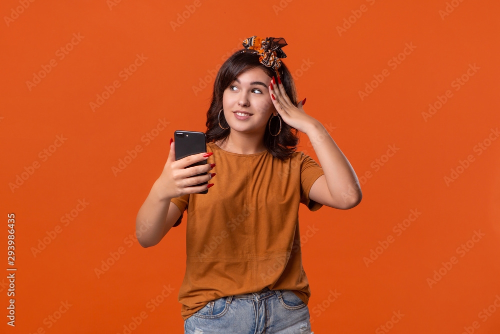 Pretty brunette woman in a t-shirt and beautiful headband making selfie with spartphone standing isolated over orange background. Mobile photography Pretty brunette woman in a t-shirt and beautiful headband making selfie with spartphone standing isolated over orange background. Mobile photography