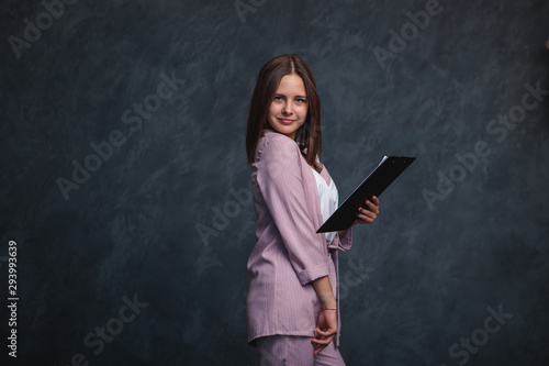 Business woman in a light suit and a black folder in her hands on a dark background stands and looks at the camera, smiles. Portrait of a business girl on a gray background, space for text.
