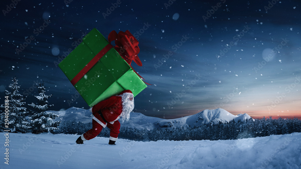 Santa Claus carrying huge christmas gift at night with copy space - obrazy, fototapety, plakaty 