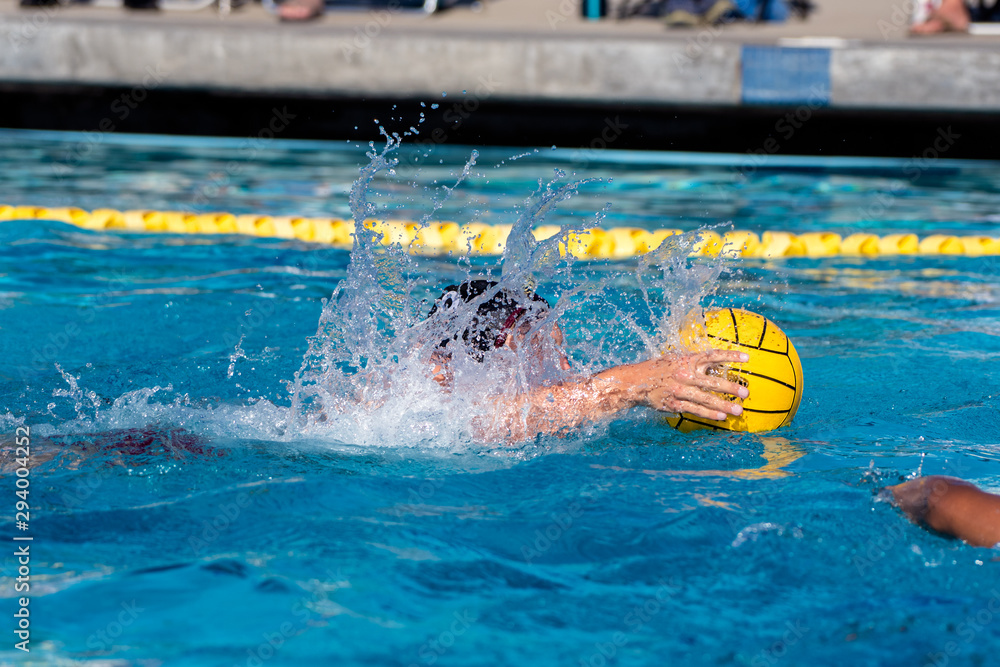 Varsity water polo player in black cap swimming while keeping the ball within his reach in the pool.