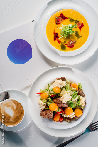 Business lunch - coffee, pumpkin cream soup and fresh vegetable salad with grilled meat in white plates on a white table in a restaurant. Close-up. Space. Flat lay. Top view