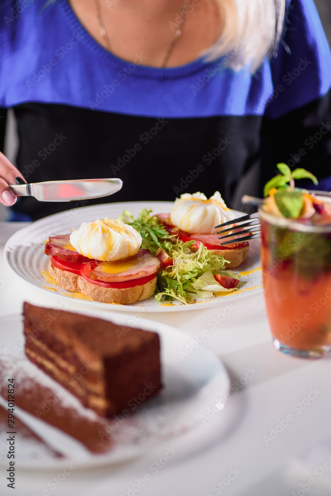 Woman eating toast with ham, tomato, poached egg and fresh salad in a restaurant. Healthy breakfast. Close-up. Space Woman eating toast with ham, tomato, poached egg and fresh salad in a restaurant. Healthy breakfast. Close-up. Space